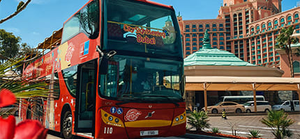Red double-decker tour bus parked on city street with historic brick buildings and covered parking structure visible in backg